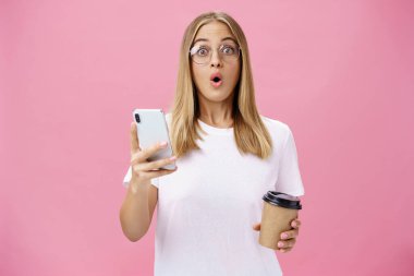 Kafedeki fincandan kahve içerken akıllı telefondan harika bir mesaj alan bir kız. Dudak büken bir arkadaşıyla birlikte haberleri paylaşıyor. Pembe duvarın üzerinde cep telefonu tutarken kameraya şaşkın şaşkın bakıyor.