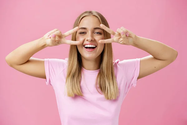 Mujer europea común alegre amistosa y pacífica con pelo rubio en camiseta casual que muestra signos de paz o discoteca sobre los ojos sonriendo alegremente a la cámara divirtiéndose contra el fondo rosa — Foto de Stock