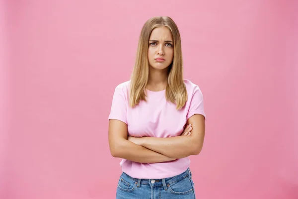 Triste envie et déçu jeune femme mignonne sombre aux cheveux blonds dans un t-shirt décontracté croisant les bras contre la poitrine dans un geste contrarié portant les lèvres et fronçant les sourcils inquiets et tristes sur un mur rose — Photo