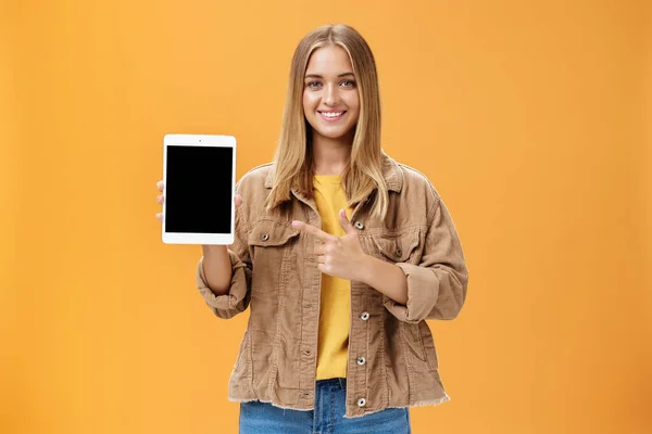 Get ready for autumn semister with new digital tablet. Portrait of charming female in corduroy jacket showing gadget screen pointing at device and smiling pleased and friendly over orange wall