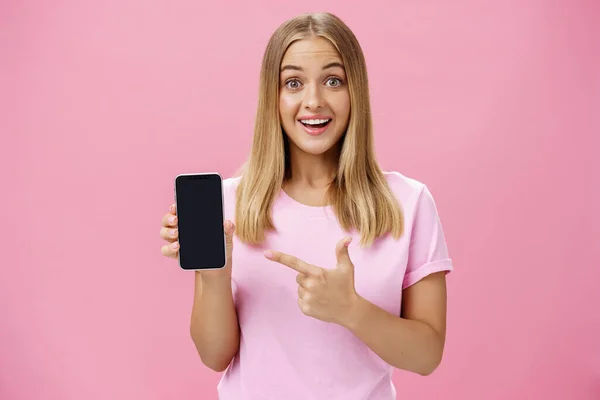 Glada attraktiv och trevlig kvinna främja cool app eller smartphone innehav mobiltelefon och pekar på enheten skärmen ler roade och imponerade stående över rosa bakgrund — Stockfoto