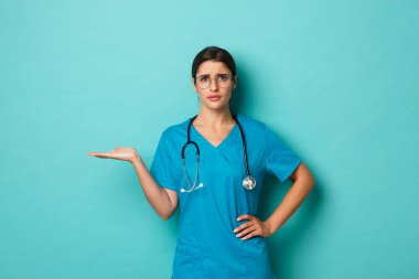 Coronavirus, salgın hastalık ve sosyalleşme konsepti. Önlüklü ve gözlüklü çekici bir kadın doktor görüntüsü, elini kaldırıyor ve kaşlarını çatıyor, bir şey anlaşılmıyor, mavi arka plan.