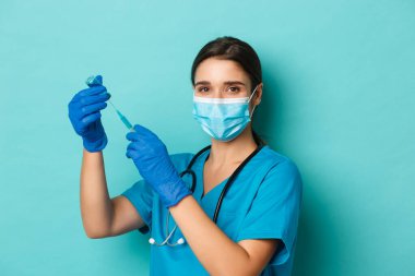 Covid-19 ve karantina kavramı. Tıbbi maskeli, eldivenli ve önlüklü kadın doktor görüntüsü. Şırıngayı koronavirüs aşısıyla dolduruyor. Mavi arka planda duruyor.