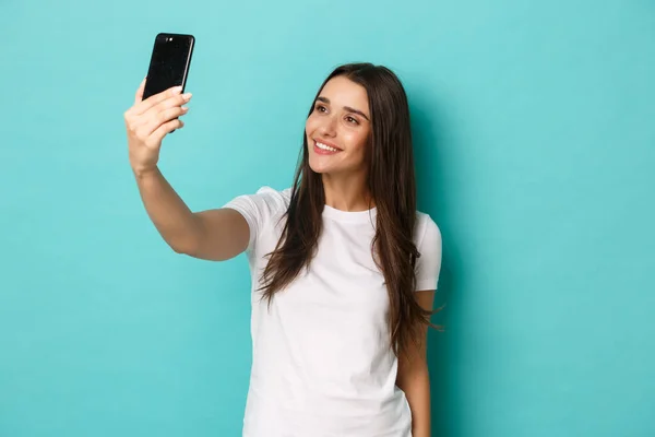 Porträtt av attraktiv brunett kvinna i vit t-shirt, ta selfie med smartphone, står över blå bakgrund — Stockfoto