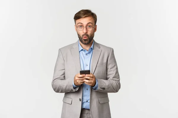 Portrait d'homme d'affaires beau avec barbe, portant costume gris et lunettes, regardant amusé après avoir lu quelque chose d'intéressant sur téléphone portable, debout sur fond blanc — Photo