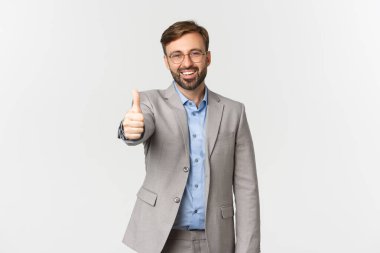 Sakallı, gözlüklü, gri takım elbiseli yakışıklı bir işadamının portresi. Başparmak kaldıran, iyi iş çıkaran, beyaz arka planda duran...