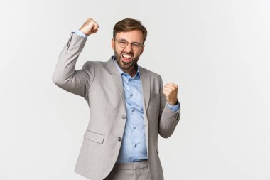 Gri takım elbiseli başarılı sakallı iş adamının portresi, hedefe ulaş ve zafere ulaş, yumruk hareketi yap ve kendine güvenerek evet de, beyaz arka planda dur.