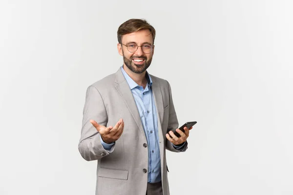 Retrato de empresário confiante com barba, vestindo óculos e terno cinza, segurando telefone celular e apontando para você satisfeito, de pé sobre fundo branco — Fotografia de Stock