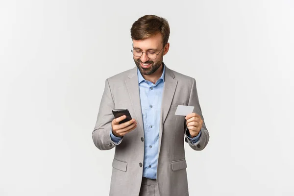 Beau homme d'affaires barbu en costume gris et lunettes, faisant du shopping en ligne, tenant carte de crédit et téléphone portable, debout sur fond blanc — Photo