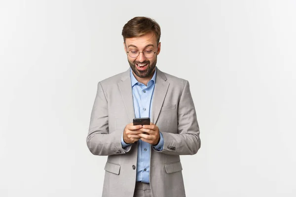 Portrait d'un bel homme d'affaires barbu, portant des lunettes et un costume gris, regardant surpris par le téléphone portable, lisant quelque chose en ligne, debout sur fond blanc — Photo