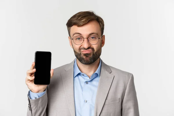 Gros plan de l'homme d'affaires sceptique en lunettes et costume gris, boudant et montrant l'écran de téléphone portable, debout sur fond blanc — Photo
