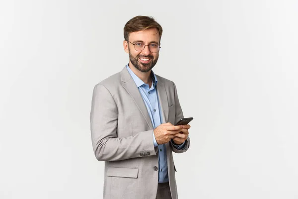 Bonito empresário com barba, vestindo óculos e terno, segurando celular e sorrindo para a câmera, em pé sobre fundo branco — Fotografia de Stock