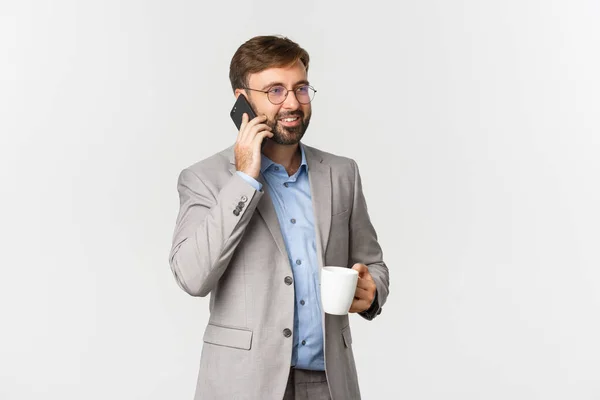 Imagen del exitoso hombre de negocios en traje gris y gafas, bebiendo café y hablando por teléfono con sonrisa complacida, de pie sobre fondo blanco —  Fotos de Stock