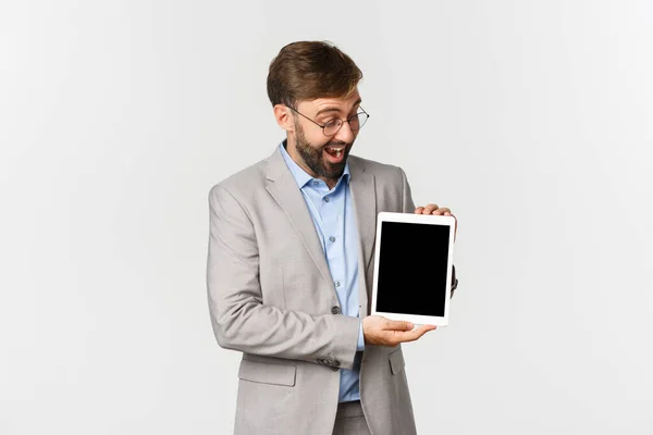 Πορτρέτο του ενθουσιασμένου επιχειρηματία με γενειάδα, φορώντας γυαλιά και γκρι κοστούμι, δείχνοντας κάτι στην ψηφιακή οθόνη tablet, κοιτάζοντας κατάπληκτος, στέκεται πάνω από το λευκό φόντο — Φωτογραφία Αρχείου