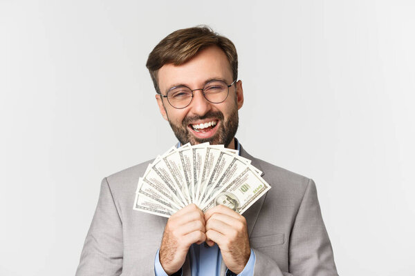 Close-up of rich and successful businessman, wearing gray suit and glasses, showing cash and laughing accomplished, standing over white background