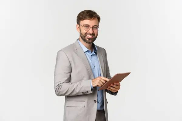 Porträtt av stilig skäggig manlig entreprenör i grå kostym och glasögon, med digital tablett och arbetar, ler glad mot kameran, står över vit bakgrund — Stockfoto