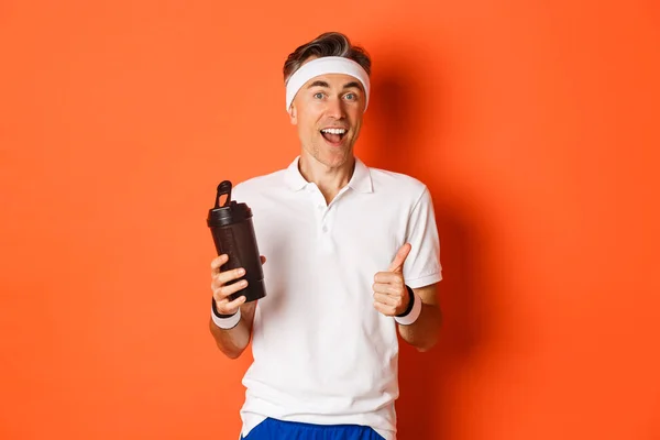 Porträt eines gesunden und aktiven Fitness-Typen mittleren Alters, der Turnuniform trägt, Daumen nach oben zeigt, Wasser trinkt und vor orangefarbenem Hintergrund steht — Stockfoto