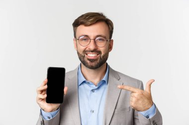 Gözlüklü ve gri takım elbiseli kendine güvenen işadamlarının yakın çekimi, cep telefonu ekranını işaret ediyor ve gururla gülümsüyor, beyaz arka planda duruyor.