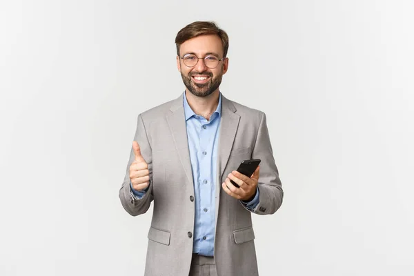 Portrait d'un bel homme d'affaires avec barbe, portant des lunettes et une combinaison grise, tenant le téléphone portable et montrant les pouces en l'approbation, souriant heureux, fond blanc — Photo