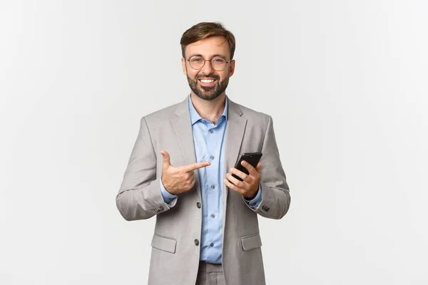 Image d'un bel entrepreneur masculin avec barbe, portant des lunettes et un costume gris, pointant vers le téléphone portable avec un sourire heureux, montrant l'application, debout sur fond blanc — Photo