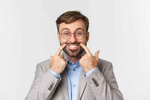 Close-up de homem de negócios em terno cinza e óculos tentando puxar um sorriso feliz, de pé sobre fundo branco — Fotografia de Stock