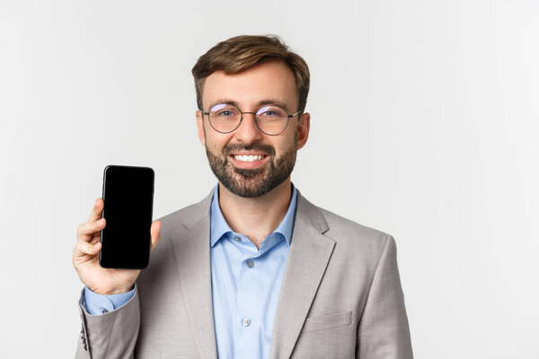 Close-up of handsome businessman in glasses and gray suit showing mobile phone screen, demonstrating an app, standing over white background