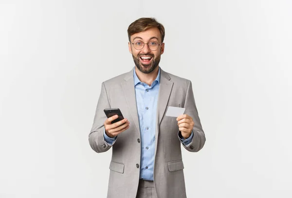 Portrait d'entrepreneur masculin barbu en costume et lunettes, l'air excité tout en faisant des achats en ligne avec téléphone mobile et carte de crédit, debout sur fond blanc — Photo