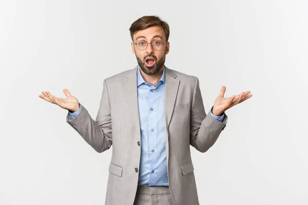 Portrait of surprised businessman with long beard, wearing grey suit and glasses, spread hands sideways and open mouth confused, standing puzzled over white background — Stock Photo, Image