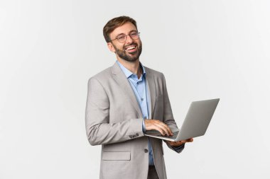Gri takım elbiseli ve gözlüklü başarılı iş adamının portresi, dizüstü bilgisayarla çalışıyor ve kameraya mutlu bir şekilde gülümsüyor, beyaz arka planda duruyor.