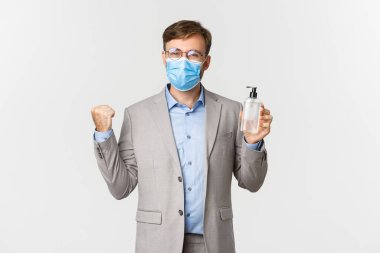 İş anlayışı, covid-19 ve sosyal mesafeler. Neşeli ve kendinden emin iş adamının görüntüsü çalışanları el dezenfektanı ya da antiseptik kullanmaya teşvik ediyor, tıbbi maske ve gri takım elbise içinde duruyorlar.