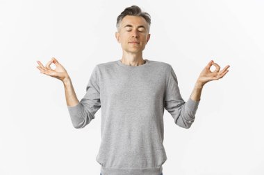 Sakin ve rahat orta yaşlı bir adamın portresi, rahatça nefes alıyor, gözleri kapalı meditasyon yapıyor ve elleri yana doğru açık, beyaz arka planda yoga yapıyor.