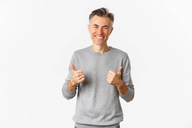 Gri saçlı, yakışıklı, orta yaşlı bir adamın portresi. Baş parmak işareti yapıyor ve gülümsüyor. Beyaz arka planda duran fikir gibi güzel bir şeyi onaylıyor.