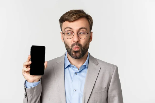 Gros plan d'un homme d'affaires excité en costume gris et lunettes, montrant l'écran de téléphone portable et regardant heureux, debout sur fond blanc — Photo