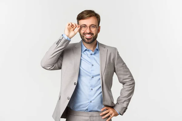 Porträtt av attraktiv framgångsrik affärsman med skägg, bär glasögon och grå kostym, ler nöjd och säker, står över vit bakgrund — Stockfoto