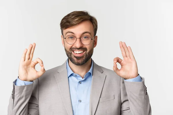Close-up de homem barbudo bonito em terno cinza e óculos, recomendar algo, mostrando sinal ok e sorrindo, aprovar boa escolha, de pé sobre fundo branco — Fotografia de Stock