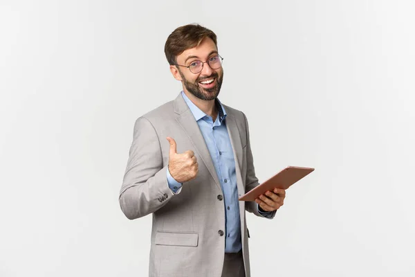 Retrato de empresário feliz em terno cinza e óculos, elogiando o bom trabalho, lendo algo no tablet digital e mostrando polegares para cima na aprovação, de pé sobre fundo branco — Fotografia de Stock