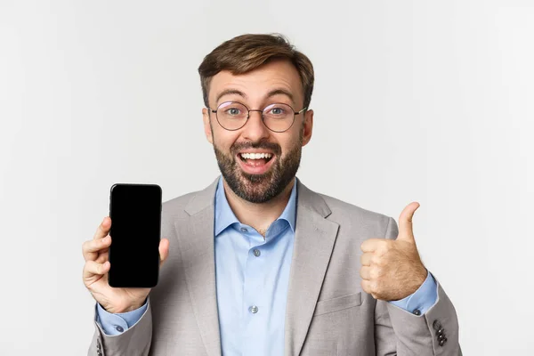 Gros plan de l'homme d'affaires excité en costume gris et lunettes, montrant pouces vers le haut et l'écran de téléphone mobile, montrant quelque chose d'étonnant, debout sur fond blanc — Photo