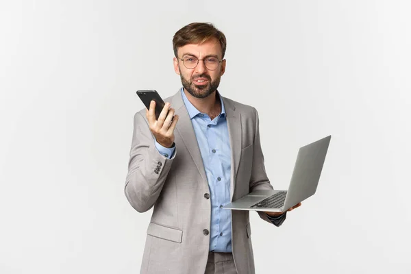 Image d'un homme d'affaires confus en costume gris et lunettes, recevoir de mauvaises nouvelles lors d'un appel téléphonique, tenant ordinateur portable et smartphone, regardant la caméra perplexe, fond blanc — Photo