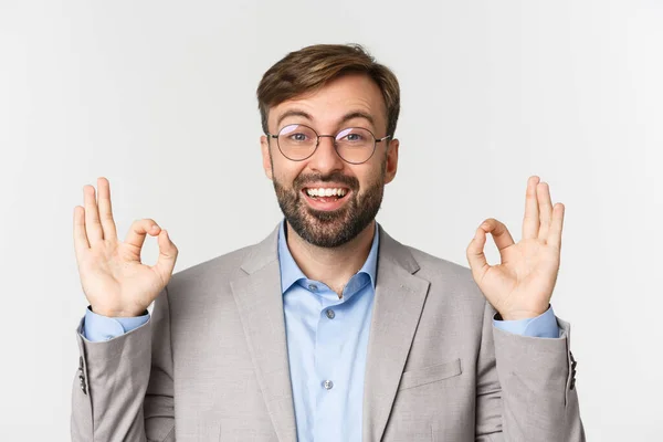Close-up de empresário confiante bonito, vestindo óculos e camisa cinza, mostrando sinal ok e sorrindo, aprovar algo bom, de pé sobre fundo branco — Fotografia de Stock