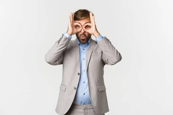 Porträt eines lustigen bärtigen Geschäftsmannes im grauen Anzug, der eine Fingerbrille aufsetzt und durch sie auf etwas Überraschendes blickt, das vor weißem Hintergrund steht — Stockfoto