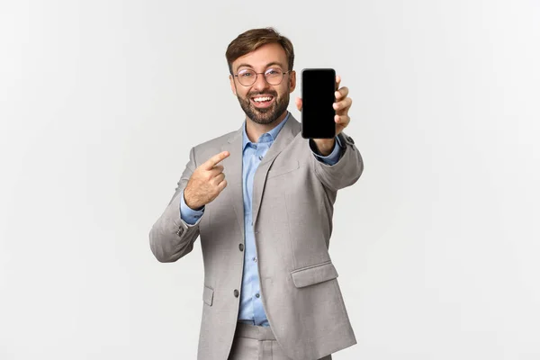 Obrázek vzrušeného bělošského obchodníka s vousy, v brýlích a šedém obleku, něco na obrazovce chytrého telefonu, stojící nad bílým pozadím — Stock fotografie