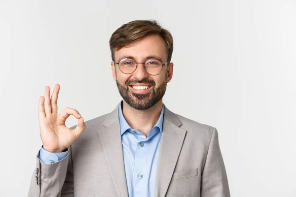 Close-up de belo empresário confiante, vestindo óculos e camisa cinza, mostrando sinal ok e sorrindo, aprovar algo, de pé sobre fundo branco — Fotografia de Stock