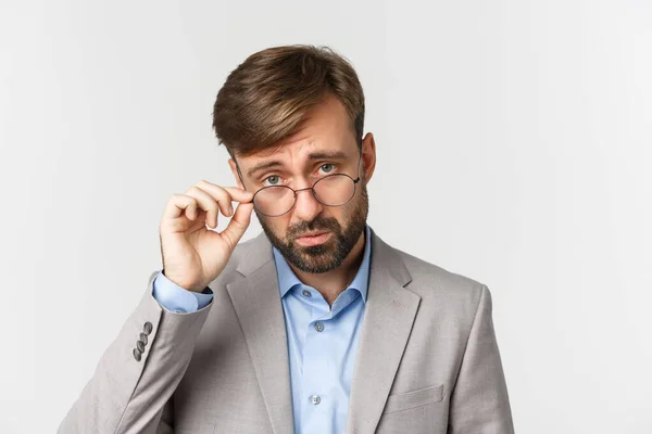 Close-up of skeptical bearded man, looking from under glasses and frowning disappointed, standing in gray suit over white background — Stock Photo, Image