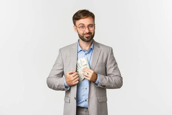 Retrato de empresario barbudo seguro y exitoso, vestido con traje gris y gafas, poniendo dinero en el bolsillo interior, sonriendo complacido, de pie sobre fondo blanco — Foto de Stock