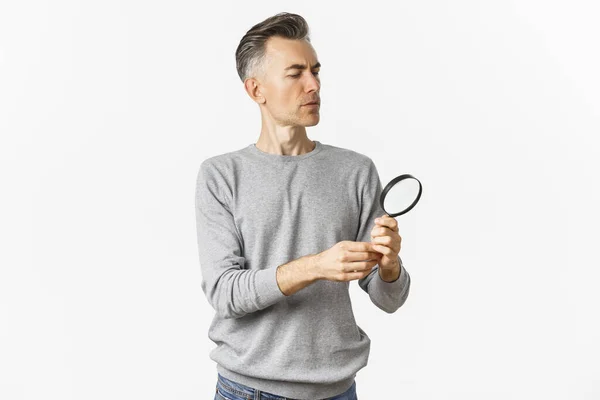 Retrato de homem pensativo de meia-idade olhando através de lupa, à procura de algo ou leitura, de pé sobre fundo branco — Fotografia de Stock
