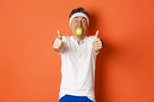 Bild des lustigen Typen mittleren Alters in Fitnessbekleidung, den Apfel im Mund haltend, Daumen hoch, auf orangefarbenem Hintergrund stehend — Stockfoto