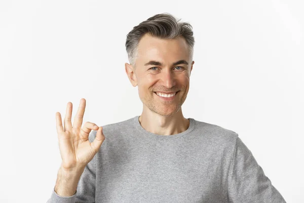 Primer plano de hombre de mediana edad guapo, mostrando signo bien y sonriendo, garantía de calidad, asegurar que todo lo bueno, de pie sobre fondo blanco — Foto de Stock