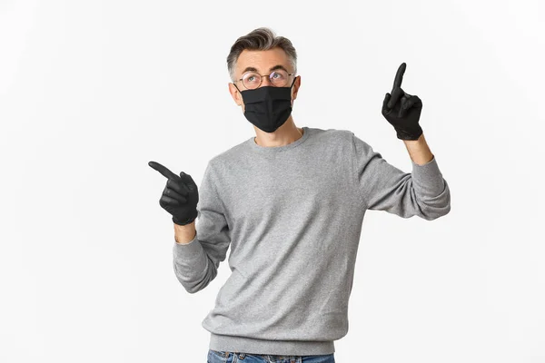Концепция коронавируса, образа жизни и карантина. Изображение мужчины средних лет с седыми волосами, в черной медицинской маске, перчатках и очках, указывающих пальцами в сторону, показывающих пространство для копирования — стоковое фото