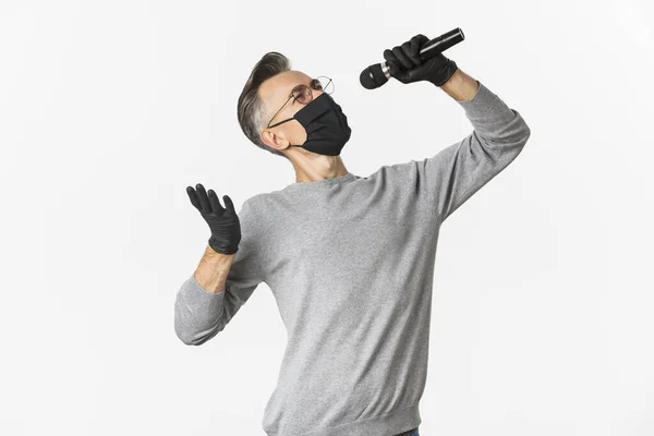 Konzept Coronavirus, Lebensstil und Quarantäne. Porträt eines attraktiven Mannes mittleren Alters in schwarzer medizinischer Maske und Handschuhen, singend in Mikrofon, vor weißem Hintergrund stehend — Stockfoto