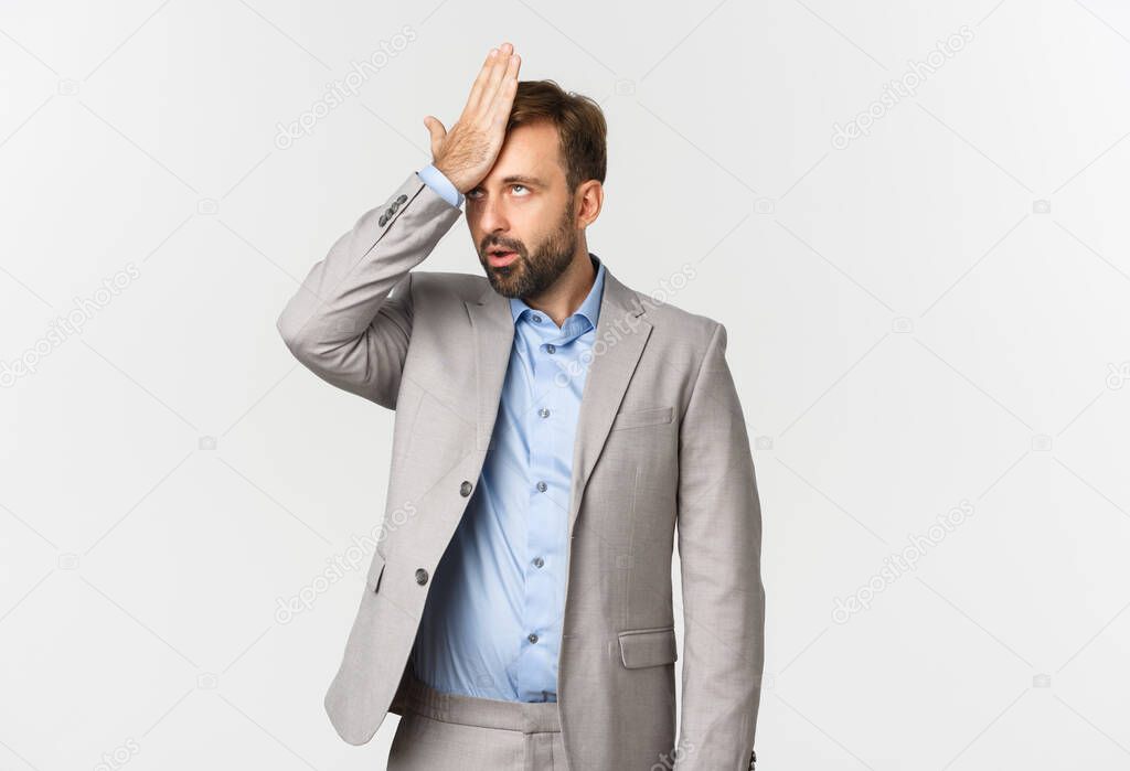 Portrait of annoyed male entrepreneur in grey suit, slap his forehead and roll eyes, bothered by something stupid, making facepalm, standing over white background